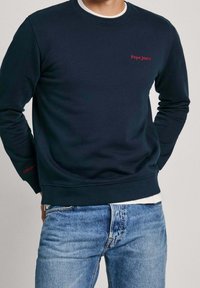 Sudadera de color navy hecha de una tela suave, con cuello redondo, puños acanalados y un logo bordado en rojo en el pecho y la manga. Combinada con jeans azules.