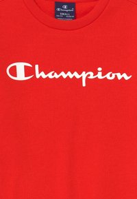 Czerwona bawełniana koszulka z białym logo "Champion" w stylu pisma odręcznego, z okrągłym dekoltem i gładką fakturą. Rozmiar: mały.