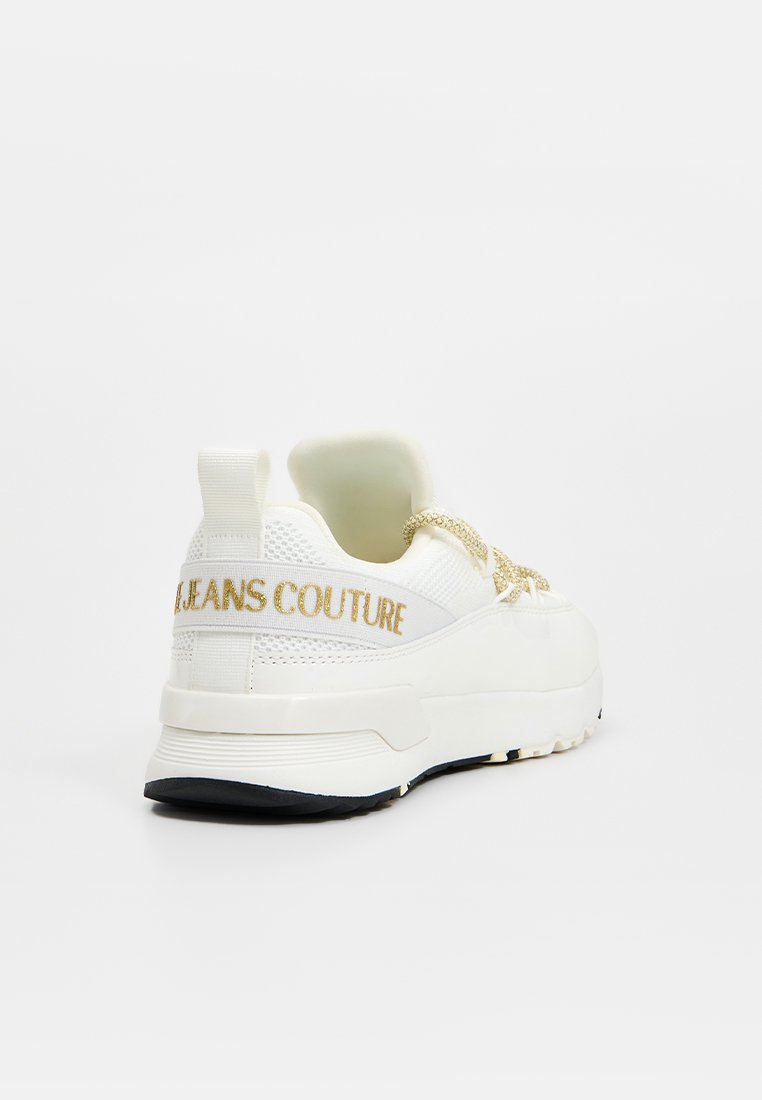 Zapatilla blanca con suela gruesa, cordones en tono dorado y "JEANS COUTURE" bordado en dorado en la correa del talón, mostrada desde la parte trasera.