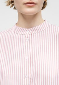 Κοντινό πλάνο ενός ατόμου που φοράει ροζ και λευκό κατακόρυφα ριγέ πουκάμισο με στρογγυλή γιακά και χρυσούς σκουλαρικια κρίκους.