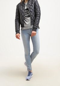 Chaqueta bomber acolchada negra sobre un sudadera gris con texto gráfico, jeans ajustados azul claro y zapatillas deportivas azul claro.