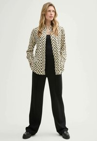Gestreifte Bluse in Beige und Schwarz, kombiniert mit schwarzen weiten Hosen. Die Bluse hat einen Kragen und eine durchgehende Knopfleiste. Glattes Material.
