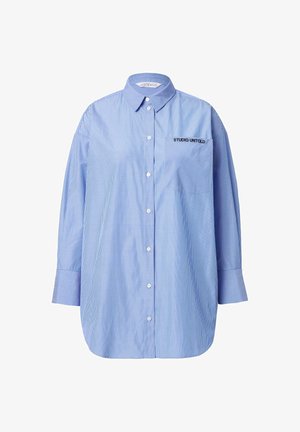Chemise bleue à rayures avec une poche arborant le texte "STUDIO UNTOLD", col classique et manches longues.