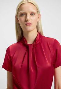 Blusa rossa di raso con colletto alto, maniche corte e dettaglio annodato al collo. Tessuto liscio con finitura lucida.