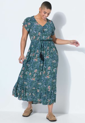 Kvinde med kort hår iført en grøn midikjole med blomsterprint og snøre i taljen samt beige slip-on sko, stående op ad en hvid væg.