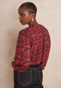 Blusa rossa e blu navy con motivo a fantasia, maniche a sbuffo e scollo a barchetta, abbinata a jeans di denim scuro. Design floreale testurizzato su tutta la superficie.