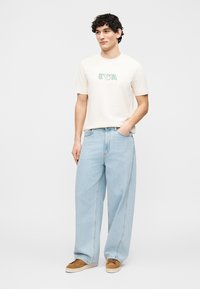 Νεαρός άνδρας που φοράει λευκό μπλουζάκι με το λογότυπο «EVA», ανοιχτόχρωμα φαρδιά τζιν και καφέ αθλητικά παπούτσια, στέκεται με το ένα χέρι στην τσέπη.