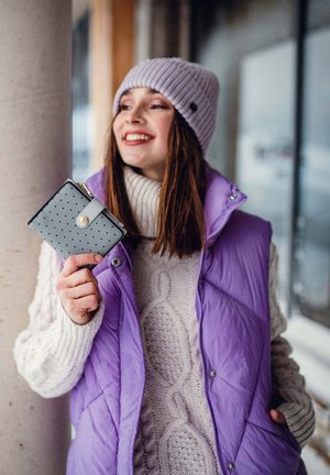 Light purple quilted vest over a cream cable-knit sweater, holding a grey wallet with perforated pattern and a gold button closure.