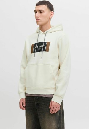Junger Mann, der einen cremefarbenen Hoodie mit einem schwarzen und braunen Jack & Jones-Logo trägt, kombiniert mit dunkel verwaschenen Jeans, steht vor einem einfarbigen Hintergrund.
