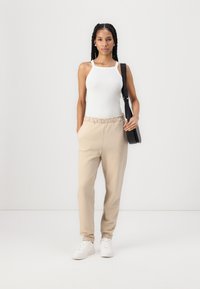 Beige sweatpants met elastische tailleband en zijvakken, gecombineerd met een aansluitend wit tanktop. Model draagt witte sneakers en heeft een zwarte tas bij zich.