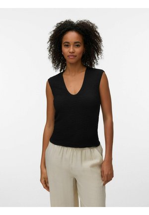 Kobieta o kręconych włosach ubrana w czarny bezrękawnik z fakturą i beżowe luźne spodnie, stojąca na jednolitym jasnym tle.