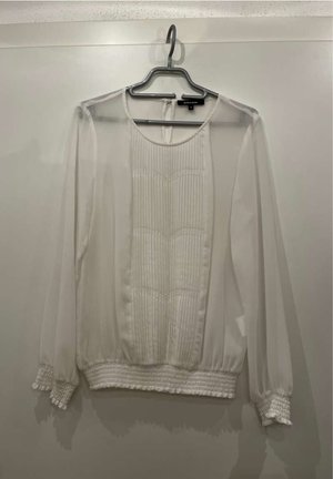 Blusa blanca de mangas largas con frente plisado, mangas transparentes, cintura elástica y puños fruncidos. Tejido texturizado con un acabado suave.