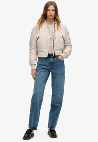 Kvinne står med den ene hånden i lommen iført en beige bomberjakke, lys grå skjorte, blå jeans, svart belte og svarte sko på hvit bakgrunn.
