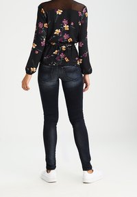 Blouse noire à motifs floraux avec manches longues et empiècement en mesh transparent, associée à un jean skinny bleu foncé et des baskets blanches.