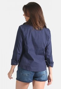 Chemise col bleu marine avec des manches reposées, fabriquée dans un tissu lisse, avec une coupe décontractée et un empiècement dans le dos. Portée avec un short en jean.
