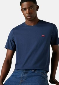 Młody mężczyzna w gładkiej granatowej koszulce Levi's i niebieskich dżinsach, pozujący na białym tle.