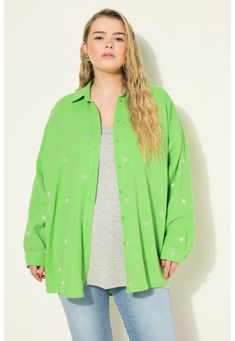 Zelená košile s knoflíky, s embosovanými stříbrnými hvězdnými vzory, texturovaná tkanina, uvolněný střih, nošena přes šedé tričko, spárovaná se světle modrými džíny.