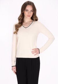 Maglione crema a V con rifiniture nere sul collo e sui polsini, maniche lunghe, texture a coste e fit aderente. Indossato con pantaloni neri.