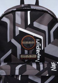 Schwarzer Rucksack mit geometrischen grauen und weißen Mustern. Verfügt über zwei Stoffaufnäher mit dem "Napapijri"-Logo und einem runden Emblem. Zipper-Detail sichtbar.