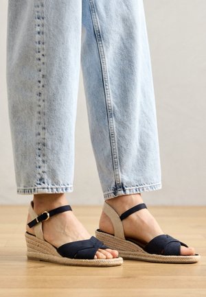 Αναδιπλωμένα σανδάλια με ναυτική ύφασμα και μπεζ λεπτομέρειες, με πλεχτή σόλα από τζούτ και ρυθμιζόμενες λωρίδες στον αστράγαλο. Φοριούνται με ανοιχτό μπλε τζιν.