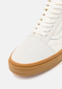 Zbliżenie na białe zamszowe sneakersy z białymi sznurowadłami i gumową podeszwą na jasnym tle.