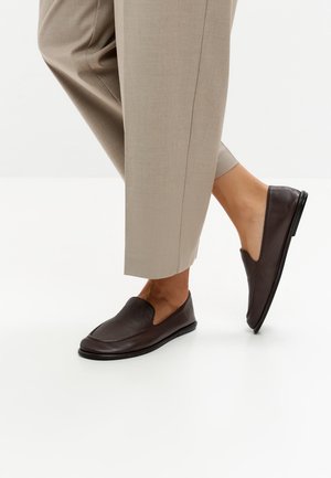 Person iført beige korte bukser og mørkebrune læder slip-on loafers, den ene fod let løftet, på hvid baggrund.