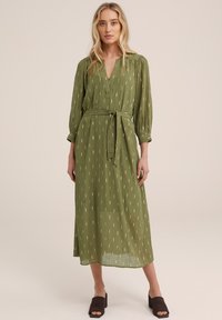 Rochie lungă verde, cu decolteu în V, talie legată și mâneci vaporoase. Prezintă un model subtil de linii verticale în nuanțe mai deschise.