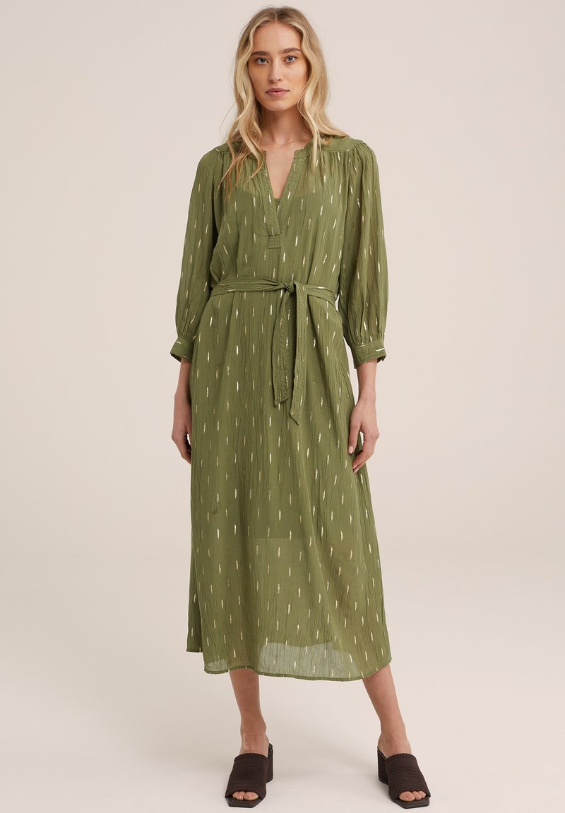 Rochie lungă verde, cu decolteu în V, talie legată și mâneci vaporoase. Prezintă un model subtil de linii verticale în nuanțe mai deschise.