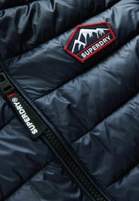 Chaleco acolchado azul marino con textura de acolchado, que presenta una insignia de montaña bordada en rojo y blanco y una cremallera negra con la marca "SUPERDRY".