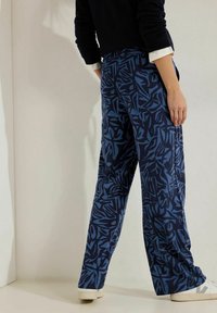 Marinblå byxor med ett abstrakt blått blommönster, tillverkade av mjukt tyg med en avslappnad passform och vida ben, som är kombinerade med ljusa skor.