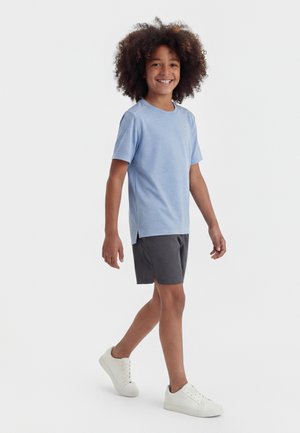 Smilende barn med krøllet hår, der går og har en lyseblå kortærmet skjorte, mørke shorts og hvide sneakers på en ensfarvet baggrund.