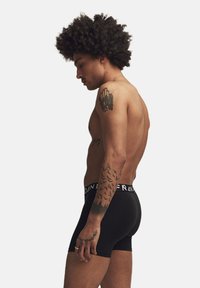 Svart boxerkalsonger med logoband, bärs av en modell med lockigt hår och tatueringar, visad i profil mot en enkel bakgrund.