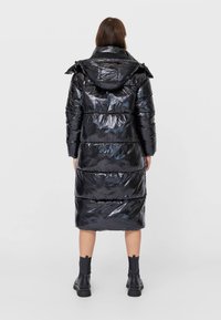 Černý péřový kabát s lesklým povrchem, po kolena, s kapucí a prošívaným designem. Nosit s černými kotníkovými botami, viditelná textura a švy.