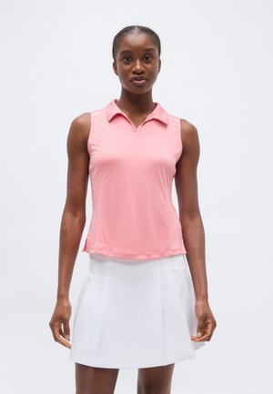 Frau trägt ein ärmelloses rosa Poloshirt und weißen Plisseerock, steht vor einem schlichten weißen Hintergrund.