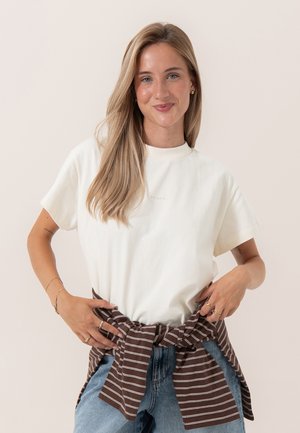 Ung kvinde med langt blondt hår iført en hvid t-shirt og blå jeans, der binder en brunstribet sweater om taljen og smiler.