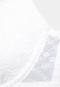 Soutien-gorge en dentelle blanche avec texture florale, panneaux transparents et détails de couture subtils. Finition lisse, bords arrondis et design sans coutures.