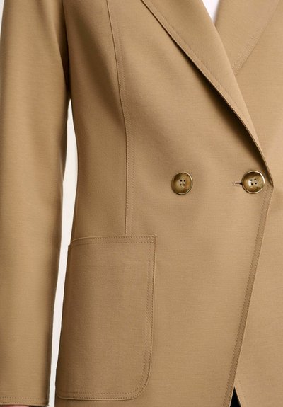 Double-breasted beige blazer with a smooth texture, gold buttons, and a front pocket. Features sharp lapels and tailored fit.
