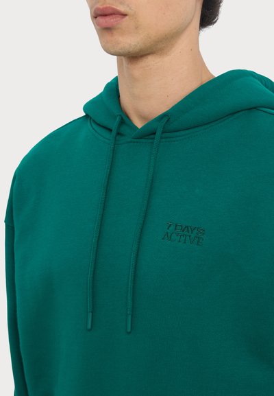 Green hoodie made of cotton, featuring a drawstring hood and a small embroidered logo "7 DAYS ACTIVE" on the chest. Soft texture.