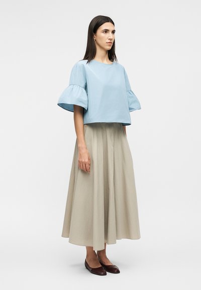 Femme debout avec des cheveux noirs raides, portant un haut bleu clair à manches courtes avec des volants, une longue jupe beige et des chaussures plates marron foncé.