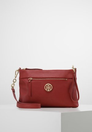 Rode leren crossbodytas met gouden rits en logo-embleem, afneembare band en pols lus op een witte ondergrond met een effen achtergrond.