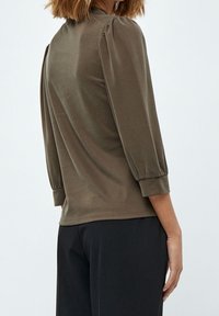 Brun, åtsittande topp med trekvartslånga puffärmar, tillverkad av mjukt material. Har en rund halsringning och rynkade detaljer vid axlarna.