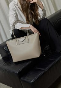 Vrouw in een wit shirt en zwarte broek zit op een zwart leren bank en houdt een grote beige handtas met dubbele handvatten vast.