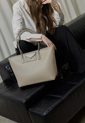 Una donna con camicia bianca e pantaloni neri è seduta su un divano in pelle nera, mentre tiene una grande borsa beige con doppi manici.