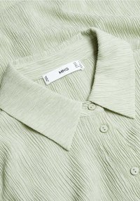 Chemise en tissu texturé vert clair avec un col pointu et trois boutons visibles, affichant une étiquette de marque blanche indiquant MNG.