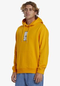 Yellow cotton hoodie with a front pocket, adjustable drawstring hood, and a black graphic logo on the chest. Ribbed cuffs and hem.