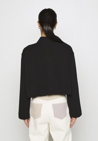 Frau mit dunklem Haar im Pferdeschwanz, die eine schwarze cropped Jacke und weiße Hosen mit zwei unterschiedlich farbigen Gesäßtaschen trägt, mit dem Rücken zugewandt.