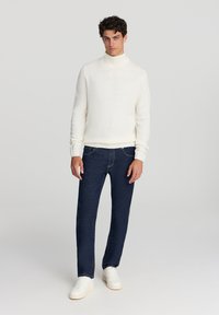 Maglione girocollo crema con polsini a costine, abbinato a jeans denim blu scuro. Il modello indossa scarpe da ginnastica bianche, mentre si trova di fronte a uno sfondo neutro.