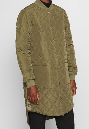 Veste longue matelassée vert olive avec boutons-pression, grandes poches avant, poignets côtelés et col côtelé, portée sur un pantalon noir.