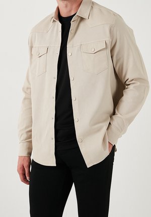 Uomo che indossa una camicia beige a maniche lunghe con bottoni sopra una t-shirt nera e pantaloni neri, in piedi con una mano in tasca.