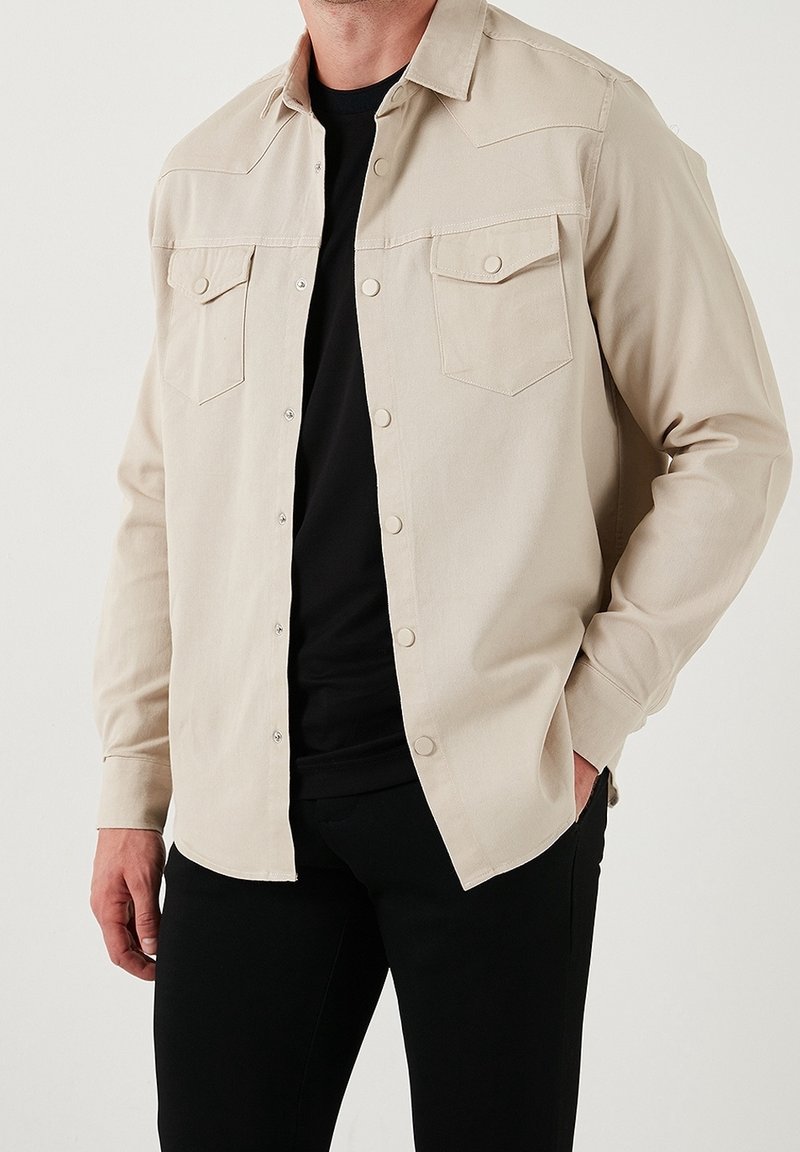 Homme portant une chemise beige à manches longues boutonnée sur un t-shirt noir et un pantalon noir, debout avec une main dans la poche.